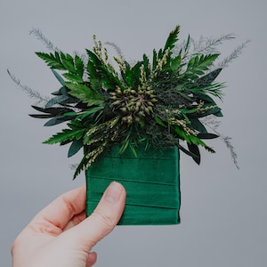 May include: A small, square, green velvet box with a variety of green foliage and small green berries arranged on top.