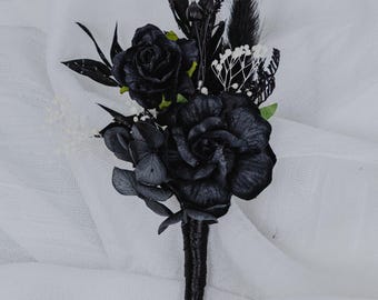 May include: A black floral boutonniere featuring two large roses, smaller flowers, and dark green leaves. The stems are wrapped in a dark, textured material. The arrangement includes white accents and feathery elements, set against a white backdrop.