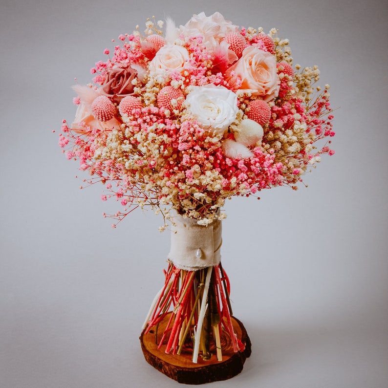 May include: A round bouquet of pink and white flowers, including roses and baby's breath. The bouquet is wrapped with a beige fabric and tied with red and white sticks, resting on a wooden base. The flowers are in various shades of pink and white.