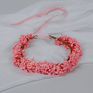 May include: A floral crown made of small, pale pink flowers. The crown is circular, with a thin, green stem base. A pink ribbon is attached for tying. Ideal for weddings or special events.