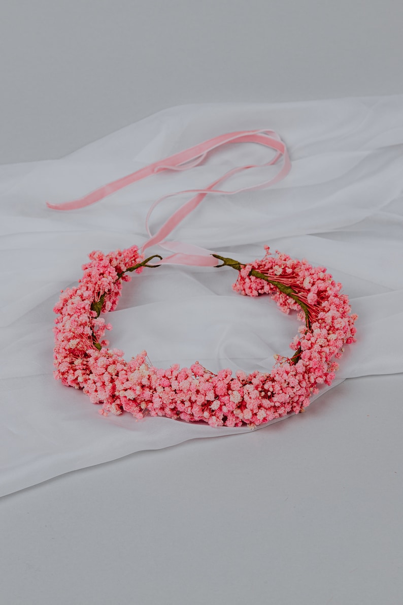 May include: A floral crown made of small, pale pink flowers. The crown is circular, with a thin, green stem base. A pink ribbon is attached for tying. Ideal for weddings or special events.