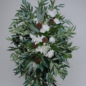 May include: A large floral arrangement featuring white flowers, pinecones, and various green leaves. The arrangement is displayed on a black stand, creating a natural and elegant display. The leaves are a mix of dark and light green.
