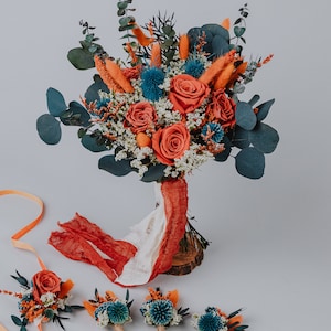 May include: A bridal bouquet featuring orange roses, blue thistle, and dried grasses, tied with a red and white fabric. Four matching buttonholes are laid out on a white surface.