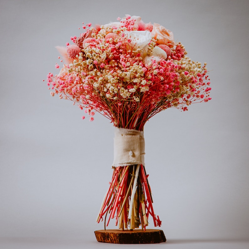May include: A wedding bouquet with a mix of pink, white, and peach flowers. The bouquet is tied with a beige fabric and rests on a small wooden base. The stems are a vibrant red.