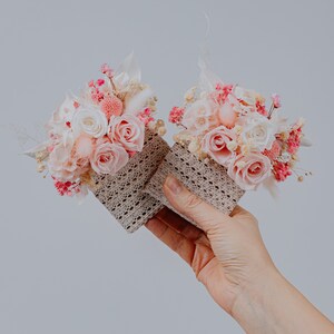 May include: Two small floral arrangements with pink and white roses, baby's breath, and dried flowers. The flowers are in square, open-weave containers. The colour scheme is soft pinks, whites, and beige.