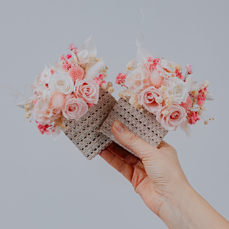 May include: Two small floral arrangements with pink and white roses, baby's breath, and dried flowers. The flowers are in square, open-weave containers. The colour scheme is soft pinks, whites, and beige.