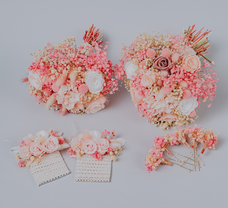 May include: A selection of pink and white floral arrangements, including two bouquets, two corsages, and a set of hair pins. The bouquets feature roses, baby's breath, and other delicate flowers. The corsages are attached to lace. The hair pins are adorned with small flowers.