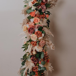 May include: A floral arrangement with a mix of peach, orange, and pink roses, greenery, and dried pampas grass.