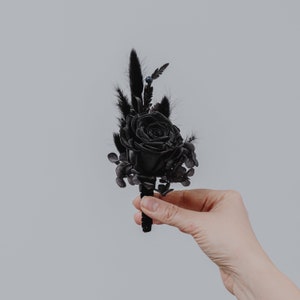 May include: A black rose buttonhole with black feathers and greenery. The buttonhole is attached to a black pin.
