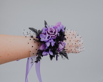 May include: A purple and white floral wrist corsage with a white polka dot mesh wristband and a purple satin ribbon.