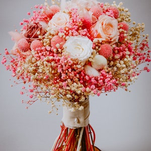 May include: A colourful bouquet of pink and white roses, baby's breath, and other flowers. The bouquet is wrapped in neutral fabric and tied with colourful stems, presented on a wooden base. A beautiful floral arrangement.