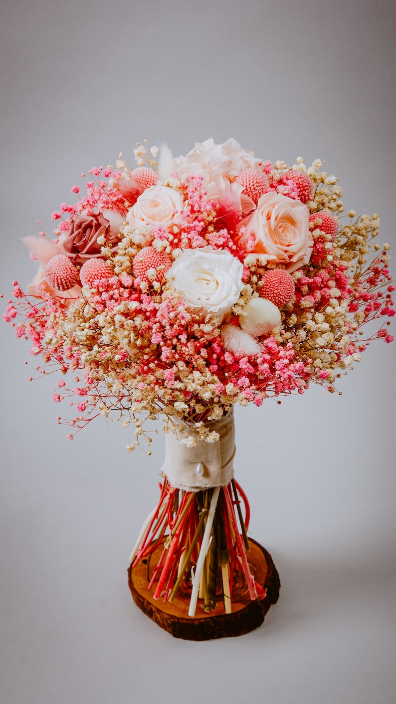 May include: A colourful bouquet of pink and white roses, baby's breath, and other flowers. The bouquet is wrapped in neutral fabric and tied with colourful stems, presented on a wooden base. A beautiful floral arrangement.