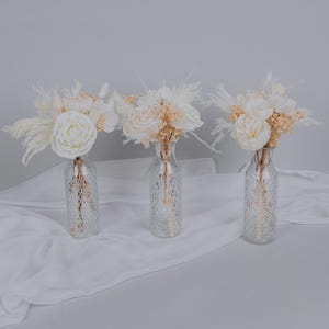 Peut inclure: Trois bouquets de fleurs séchées blanches dans des vases en verre transparent à surface texturée. Les bouquets sont disposés sur une surface en tissu blanc.