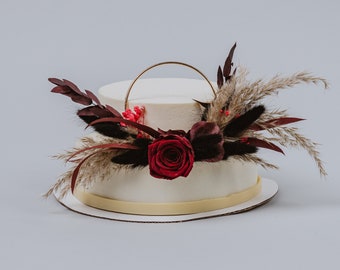 May include: A white two-tiered cake decorated with a gold ring and burgundy and red dried flowers. The cake is on a white cake stand.