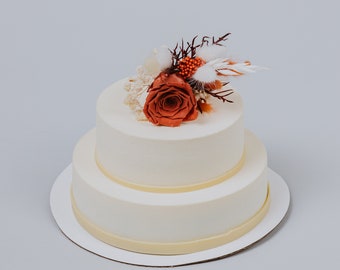 May include: A two-tiered white wedding cake decorated with a single orange rose and dried flowers. The cake is on a white cake stand.