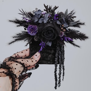 May include: A black velvet box containing a black rose and purple flowers, surrounded by black feathers. The box is being held by a black lace glove.