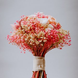 May include: A dried flower bouquet in pink, cream, and red hues. The arrangement includes small, delicate flowers and larger, textured blooms. The stems are bound with beige fabric and a pearl button, set on a wooden base. The bouquet is a charming decorative item.