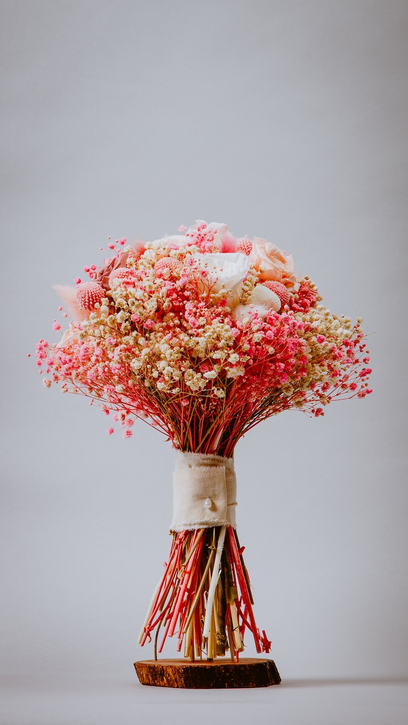 May include: A dried flower bouquet in pink, cream, and red hues. The arrangement includes small, delicate flowers and larger, textured blooms. The stems are bound with beige fabric and a pearl button, set on a wooden base. The bouquet is a charming decorative item.