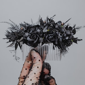 May include: A black floral hair comb with roses and dark foliage. The comb is held up against a grey background. The comb's teeth are silver. The person holding the comb is wearing a black dotted glove.