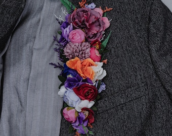 May include: A dark gray tweed blazer adorned with a vibrant floral arrangement. The flowers include shades of purple, pink, orange, and red, creating a striking contrast against the jacket's neutral tone. The lining is a light gray pinstripe.