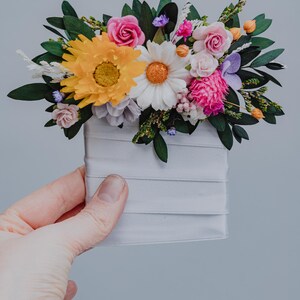 May include: A vibrant floral arrangement featuring a yellow sunflower, white daisy, and pink roses, accented with green leaves and other colorful blooms. The flowers are arranged in a white, ribbon-wrapped container.