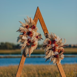 May include: A wooden A-frame arch adorned with dried pampas grass, red and white roses, and other dried flowers. The arch is positioned outdoors with a body of water and field in the background, creating a natural and rustic aesthetic.