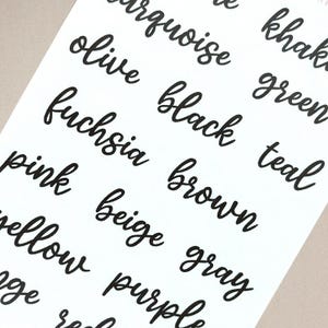 May include: A white sticker sheet featuring black text in a cursive script. The words listed include turquoise, khaki, olive, green, fuchsia, black, teal, pink, brown, beige, gray, yellow, purple, and red, arranged in multiple columns and rows.