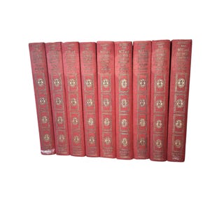 May include: A set of nine red hardcover books titled The Wit and Humor Library, featuring gold-stamped lettering and decorative circular emblems on the spines. Each book spine displays the series title, volume number, and publisher name.