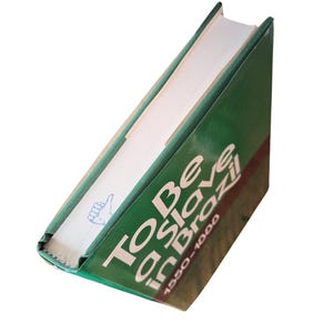 Puede incluir: Un libro de tapa dura verde titulado "To Be a Slave in Brazil 1550-1888" con texto blanco. El lomo del libro es visible, mostrando el t&iacute;tulo y una peque&ntilde;a ilustraci&oacute;n. El libro est&aacute; en &aacute;ngulo, revelando sus p&aacute;ginas.