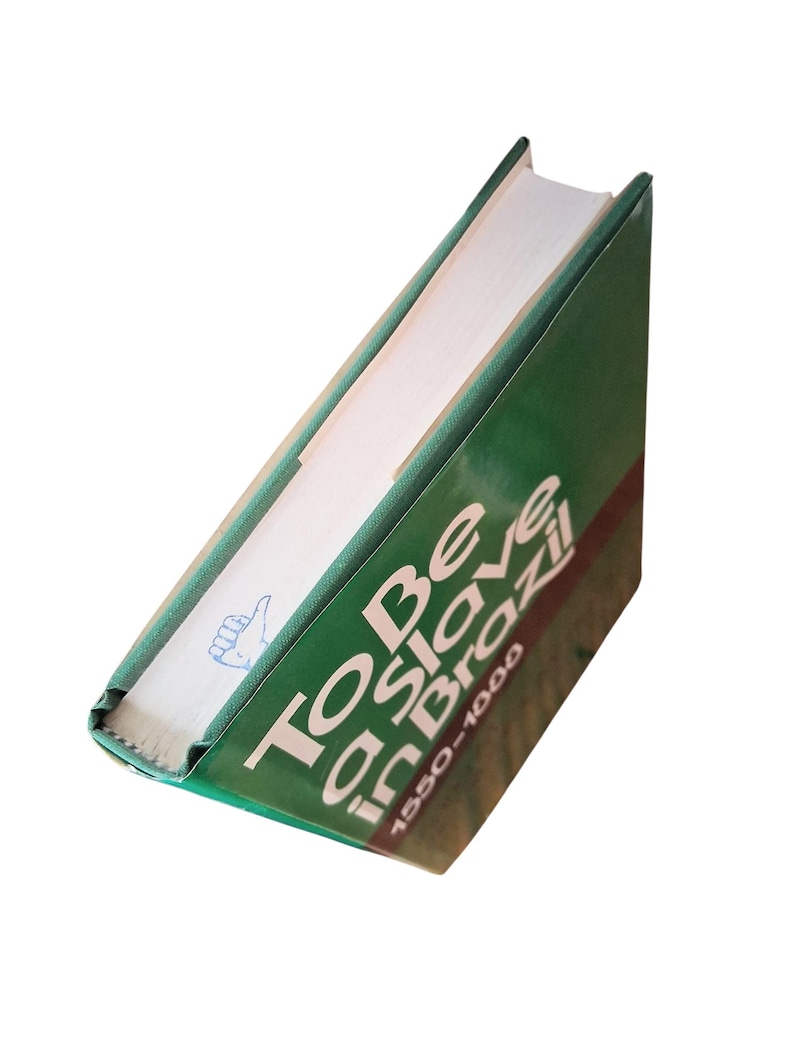 Puede incluir: Un libro de tapa dura verde titulado "To Be a Slave in Brazil 1550-1888" con texto blanco. El lomo del libro es visible, mostrando el t&iacute;tulo y una peque&ntilde;a ilustraci&oacute;n. El libro est&aacute; en &aacute;ngulo, revelando sus p&aacute;ginas.