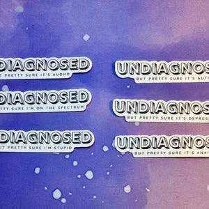 Puede incluir: Seis pegatinas blancas con texto negro sobre un fondo morado y azul. Cada pegatina dice "UNDIAGNOSED" en negrita, seguido de frases como "BUT PRETTY SURE IT'S AUTISM" o "BUT PRETTY SURE IT'S DEPRESSION".