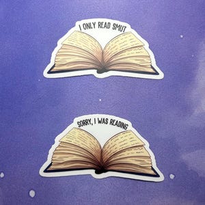 May include: Two stickers featuring open books with text. The top sticker reads "I ONLY READ SMUT." The bottom sticker says "SORRY, I WAS READING." The books have tan pages and black spines, set against a purple background.