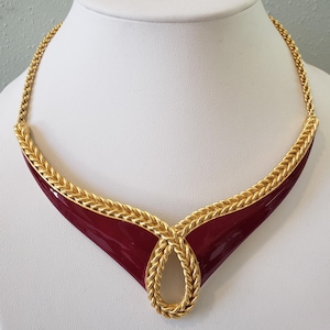 May include: A gold-toned necklace with a deep red enamel inlay. The necklace features a braided gold chain and a central teardrop design. The red enamel is a glossy, rich color, and the necklace is displayed on a white mannequin.