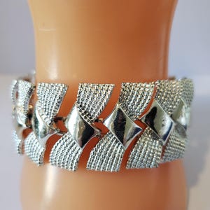 May include: Silver-toned bracelet with a textured, woven design. The bracelet features a series of connected, geometric shapes, alternating between diamond and rectangular patterns. The metal has a shiny finish, reflecting light. The bracelet is a piece of jewelry.