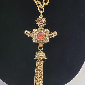 May include: Gold-toned necklace with a large pendant featuring a cross-like design. The pendant has red gemstone accents and a tassel of gold chains. The necklace is displayed against a black background.