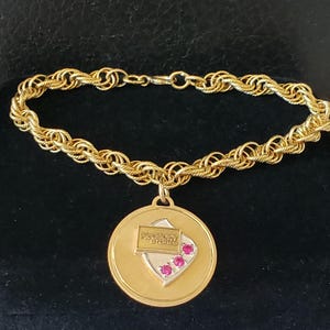 May include: A gold-toned rope chain bracelet featuring a circular pendant. The pendant displays a central shield-shaped emblem with the text McCrory Stores and three small, round, bright pink stones set in a vertical row on the right side.