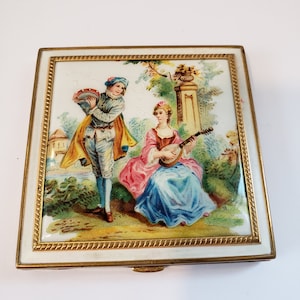 May include: A vintage, gold-trimmed, white enamel compact with a painted scene of a couple in a garden. The man is holding a tambourine and the woman is playing a lute.