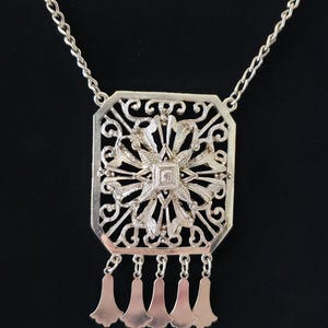 May include: Silver-toned filigree pendant necklace. The pendant has a rectangular shape with an ornate floral design and dangling bell-shaped charms. The necklace chain is a delicate link style, set against a black background.