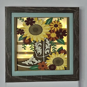 May include: A framed, layered art piece featuring a brown cowboy boot filled with sunflowers and other flowers in shades of yellow, orange, and burgundy. The artwork is illuminated with small lights, and set against a striped background. The frame is gray.