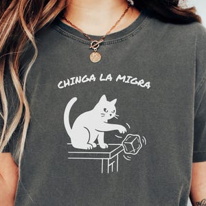 May include: Pepper-colored t-shirt with the text "CHINGA LA MIGRA" above a white graphic of a cat pushing a cube off a table. The shirt is a relaxed fit and has short sleeves.