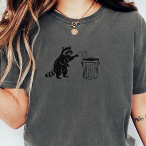 May include: Dark grey t-shirt featuring a black graphic of a raccoon throwing trash into a bin. The word "PEPPER" is printed at the bottom of the shirt. The shirt has short sleeves and a crew neck.