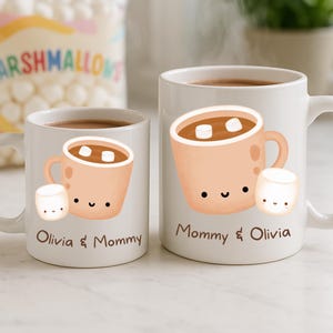 May include: Two white ceramic mugs with a hot chocolate design. Each mug features a cartoon illustration of a mug of hot chocolate with marshmallows and a smiling marshmallow. One mug says "Olivia & Mommy", the other says "Mommy & Olivia".