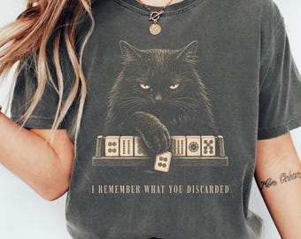 May include: A charcoal gray short-sleeve t-shirt featuring a graphic of a black cat playing mahjong with tiles on a rack. The text below the cat reads, I REMEMBER WHAT YOU DISCARDED. The shirt has a crew neckline and a relaxed, vintage-style fit.