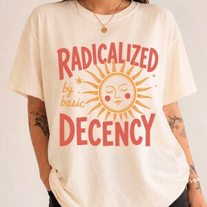 May include: An ivory short-sleeve t-shirt featuring a graphic print with the text RADICALIZED by basic DECENCY in red and orange. The design includes a central sun illustration with a face, closed eyes, and red cheeks, surrounded by rays.