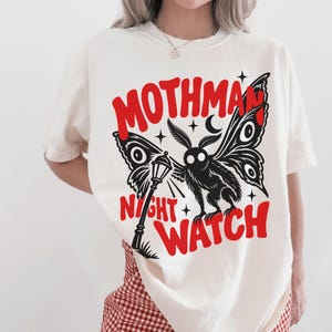 May include: An ivory t-shirt featuring a black graphic of a moth-like creature standing next to a street lamp. The text MOTHMAN NIGHT WATCH is printed in bold red letters above and below the illustration, accented by small black stars.