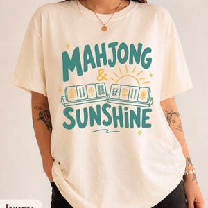 May include: An ivory short-sleeve t-shirt featuring the teal text "MAHJONG & SUNSHINE" with a yellow sun graphic and illustrations of mahjong tiles arranged in two rows. The design includes small yellow star accents and a wavy line below the text.