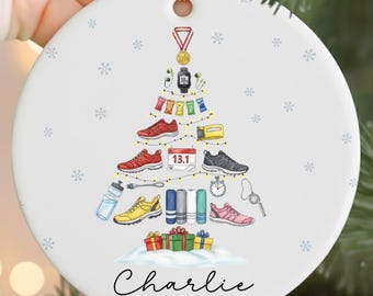 May include: White ceramic Christmas ornament with a gold string. The ornament features a Christmas tree design made of running shoes, a watch, and other running-related items. The name "Charlie" and the year "2025" are written on the ornament.
