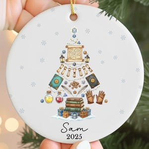 May include: A white ceramic Christmas ornament with a gold string. The ornament features a Christmas tree design made of fantasy-themed elements like books, scrolls, and dice. The name "Sam" and the year "2025" are printed at the bottom.