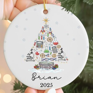 May include: A white ceramic Christmas ornament with a gold string. The ornament features a science-themed Christmas tree design with rockets, beakers, and microscopes. The name "Brian" and the year "2025" are written in black script.