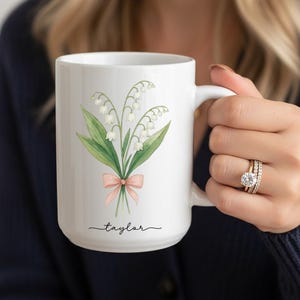 May include: White ceramic mug featuring a watercolor illustration of lily of the valley flowers tied with a pink bow. The name "Taylor" is written in a script font below the floral design. The mug has a white handle.